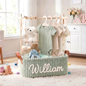 Könnte beinhalten: Ein salbeigrüner Korb aus Cord mit dem eingestickten Namen William in weißer Schreibschrift. Der Korb enthält eine hölzerne Kleiderstange mit vier Babybodys auf Holzkleiderbügeln, eine kleine Holzschaukel und verschiedene Kuscheltiere.