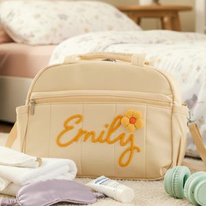 Puede incluir: Bolso cambiador crema con el nombre "Emily" bordado en amarillo, acentuado por una flor amarilla y naranja. El bolso tiene un asa superior, una correa para el hombro y un bolsillo frontal con cremallera. Junto al bolso hay toallas blancas, una m&aacute;scara de ojos lavanda, una loci&oacute;n y auriculares verde menta.