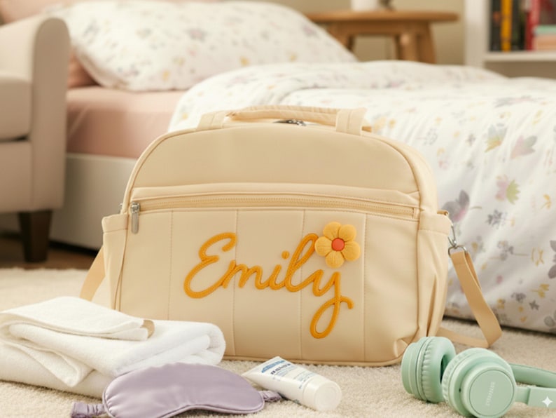 Puede incluir: Bolso cambiador crema con el nombre "Emily" bordado en amarillo, acentuado por una flor amarilla y naranja. El bolso tiene un asa superior, una correa para el hombro y un bolsillo frontal con cremallera. Junto al bolso hay toallas blancas, una m&aacute;scara de ojos lavanda, una loci&oacute;n y auriculares verde menta.