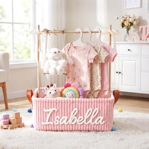 Puede incluir: Una cesta de almacenamiento de pana rosa con el nombre Isabella en letras cursivas blancas. La cesta cuenta con un marco de madera para colgar ropa con tres bodies de bebé, un conejo de peluche blanco y un juguete de arcoíris de peluche.