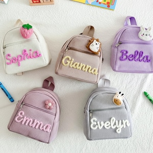 Może przedstawiać: Pięć małych plecaków sztruksowych w różnych kolorach: białym, beżowym, fioletowym, różowym i szarym. Każdy plecak ma z przodu wyszyte imię i ozdobny urok. Plecaki są przeznaczone dla dzieci.