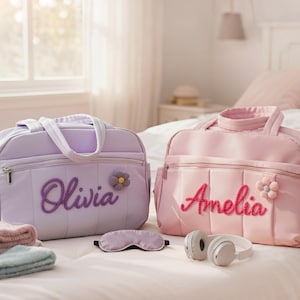 Puede incluir: Dos bolsas de beb&eacute; personalizadas, una lavanda y otra rosa, con nombres bordados y detalles de flores. Las bolsas est&aacute;n sobre una cama con toallas, una m&aacute;scara para dormir y auriculares.