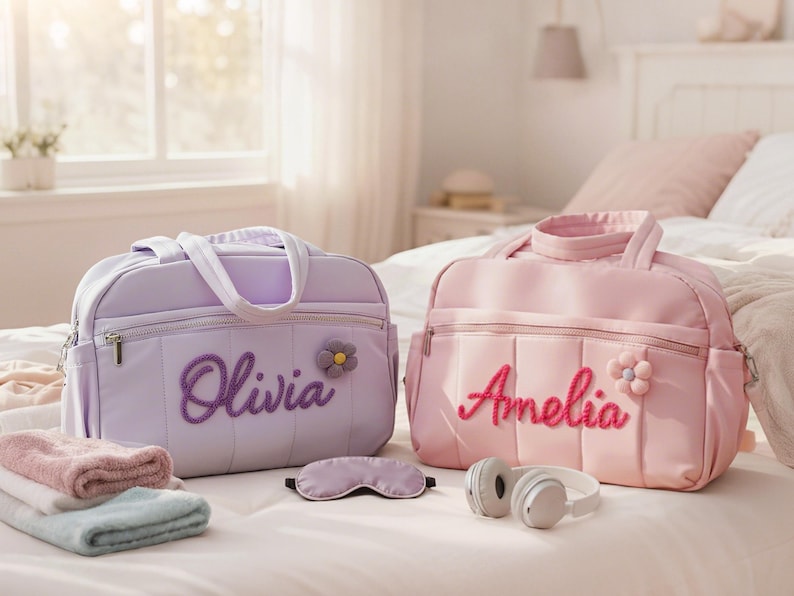 Puede incluir: Dos bolsas de beb&eacute; personalizadas, una lavanda y otra rosa, con nombres bordados y detalles de flores. Las bolsas est&aacute;n sobre una cama con toallas, una m&aacute;scara para dormir y auriculares.