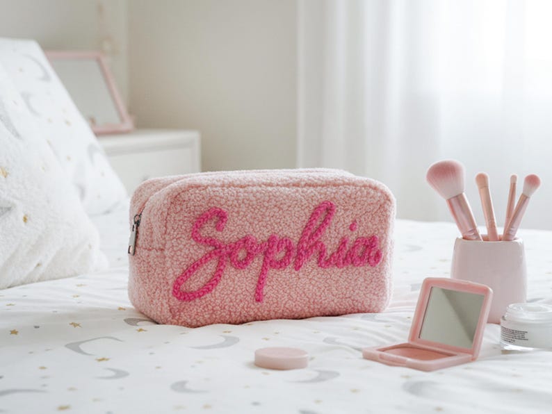 May include: A pink, textured cosmetic bag with the name "Sophia" embroidered in a darker pink script. The bag is rectangular with a zipper closure. Makeup brushes, a compact mirror, and other beauty items are nearby.