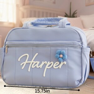 Puede incluir: Un bolso para pa&ntilde;ales azul claro, de 40 cm de ancho y 28 cm de alto, con el nombre "Harper" en escritura blanca. Cuenta con un adorno de flor azul y una cremallera plateada. Incluye una correa para el hombro y un asa superior.