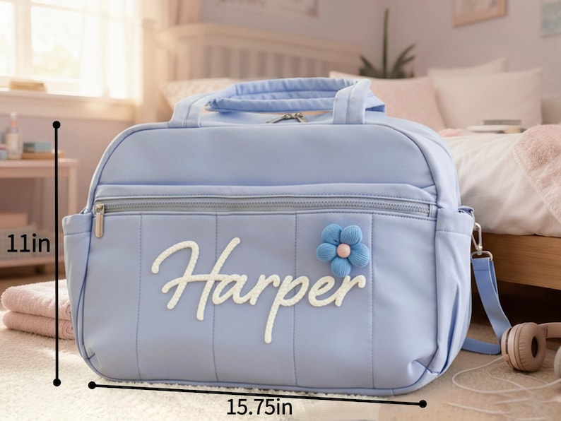 Puede incluir: Un bolso para pa&ntilde;ales azul claro, de 40 cm de ancho y 28 cm de alto, con el nombre "Harper" en escritura blanca. Cuenta con un adorno de flor azul y una cremallera plateada. Incluye una correa para el hombro y un asa superior.