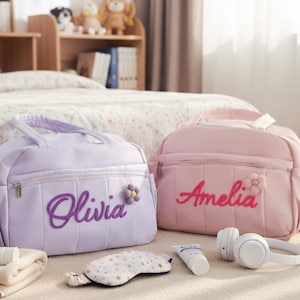 Puede incluir: Dos bolsas personalizadas, una lavanda y otra rosa, cada una con un nombre bordado en la parte delantera. La bolsa lavanda dice "Olivia" y la rosa "Amelia". Ambas bolsas tienen un adorno floral. Otros art&iacute;culos incluyen auriculares, antifaz y productos para el cuidado de la piel.