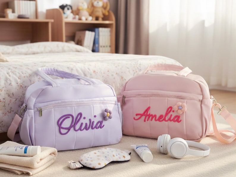 Puede incluir: Dos bolsas personalizadas, una lavanda y otra rosa, cada una con un nombre bordado en la parte delantera. La bolsa lavanda dice "Olivia" y la rosa "Amelia". Ambas bolsas tienen un adorno floral. Otros art&iacute;culos incluyen auriculares, antifaz y productos para el cuidado de la piel.