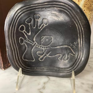 May include: A dark gray, carved stone plate with a raised border. The plate features a stylized image of an animal and a tree. The plate is displayed on a clear acrylic stand.