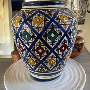 May include: A ceramic vase with a blue, white, yellow, red, and green geometric pattern. The vase has a wide base and a narrower neck. It sits on a white, textured plate. The vase is a decorative item.