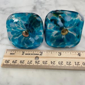 May include: Two square glass cabinet knobs featuring a mottled pattern of teal, blue, and white colors with a central brass-toned metal screw. A wooden ruler is positioned below the knobs, showing each knob is approximately 1.5 inches wide.