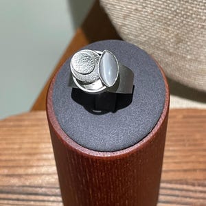 May include: A silver-toned ring featuring a wide band topped with a circular element with a textured surface, layered next to a smooth, oval-shaped gray stone. The ring is displayed on a cylindrical stand with a dark gray top.