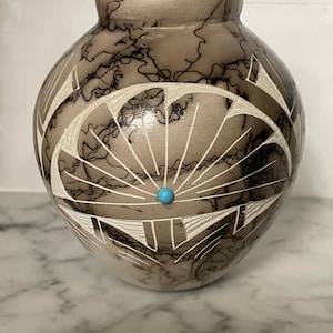 May include: A handcrafted, round pottery vase with a light brown and gray color scheme. The vase features intricate white geometric designs and a turquoise bead accent. The surface has a unique crackle pattern.