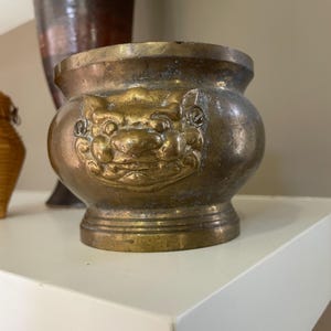 May include: A small, antique brass pot with a detailed relief of a mythical creature's face. The pot has a rounded body, a slightly flared base, and a wide opening. The brass has a warm, aged patina, suggesting its vintage. A decorative piece.
