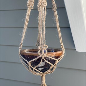 May include: A brown ceramic bowl held within a natural tan jute macrame plant hanger. The hanger features intricate knotted patterns and a central bottom tassel, suspending the glossy, dark brown glazed bowl from a single point above.