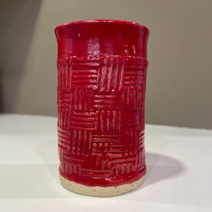 May include: A handmade, cylindrical red ceramic vase with a textured, woven pattern around the middle. The vase has a glossy finish and a natural, unglazed base. The height is approximately 5 inches.