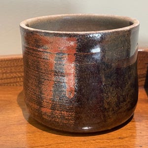 May include: A handmade ceramic mug with a cylindrical shape. The mug features a dark brown exterior with reddish-orange accents and a glossy finish. The interior is a lighter brown color. The mug sits on a wooden surface.