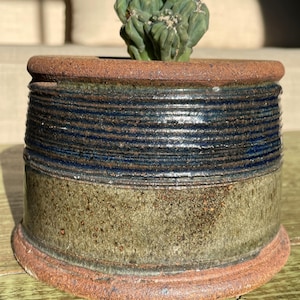 May include: A small, handcrafted ceramic pot with a cactus plant. The pot features a rustic design with a brown rim and base, and a band of dark blue and green horizontal stripes. The cactus is a vibrant green color.