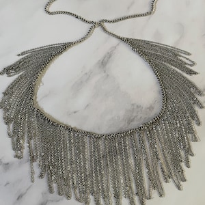 May include: Silver-toned necklace featuring a cascade of delicate chains. The necklace has a choker-style design with a series of small chains that hang down, creating a fringe effect. The necklace is made of a shiny, metallic material.