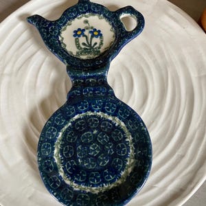 May include: A ceramic spoon rest shaped like a teapot with a dark blue and green patterned glaze. The upper section features a hand-painted floral design with two blue flowers and green leaves on a cream background, while the base has a circular pattern.