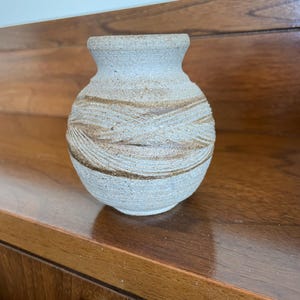 May include: A small, handcrafted ceramic vase with a rounded body and a narrow neck. The vase features a textured, light gray surface with horizontal bands of brown. The vase is displayed on a wooden surface.