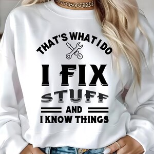 以下が含まれることがあります： 「THAT'S WHAT I DO I FIX STUFF AND I KNOW THINGS」の黒い文字がプリントされた白いスウェットシャツ。レンチとドライバーのグラフィックデザイン。クルーネックのスタイルです。