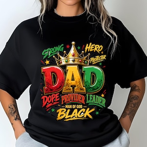 Op de afbeelding: Een zwart t-shirt met korte mouwen en een grafisch ontwerp met een gouden kroon boven het woord DAD in rood, geel en groen. De tekst bevat STRONG, HERO, PROTECTOR, DOPE, PROVIDER, LEADER, MAN OF GOD en BLACK met kleine steraccenten.