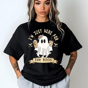 Könnte beinhalten: Schwarzes T-Shirt mit einem Halloween-Motiv. Das Design zeigt einen Cartoon-Geist mit zwei Kaffeetassen und dem Text "I'M JUST HERE FOR THE BOOS". Das Shirt hat kurze Ärmel und einen Rundhalsausschnitt.