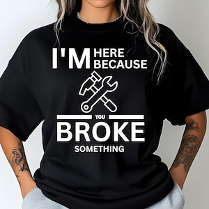 May include: Black crew neck t-shirt with the text "I'M HERE BECAUSE YOU BROKE SOMETHING" in white. A hammer and wrench graphic is centered below the text. The shirt is a casual style.