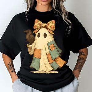 May include: Black crew neck sweatshirt featuring a whimsical ghost design. The ghost is adorned with a floral bow, patchwork details, and holds a crow. The design has a vintage, autumnal aesthetic.