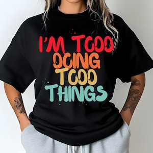 Puede incluir: Camiseta negra con el texto "I'M TOO DOING TOO THINGS" en rojo, naranja, amarillo y turquesa. La camiseta tiene cuello redondo y mangas cortas. La persona lleva un pantalón de chándal gris.
