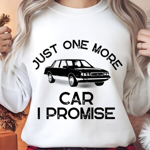 Pode incluir: Camisola branca com o texto preto "JUST ONE MORE CAR I PROMISE". Um gráfico de carro preto está centrado na frente. A camisola é usada por uma pessoa.