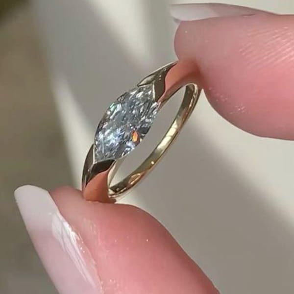 Może przedstawiać: Złoty pierścionek z diamentem o szlifie markizy. Obręcz pierścionka ma pleciony wzór po bokach, a diament jest utrzymywany na miejscu przez pazury. Pierścionek jest trzymany między dwoma palcami.
