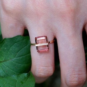 May include: A gold ring featuring a rectangular, light pink gemstone. A horizontal gold bar bisects the gemstone. The ring is set on a finger, with green leaves in the background. The ring is a piece of jewelry.