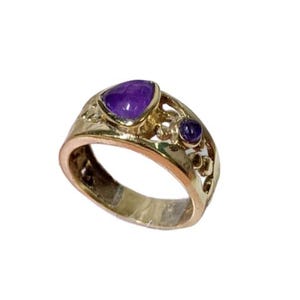 May include: A gold-toned ring featuring a large, pear-shaped purple gemstone and two smaller round purple gemstones. The band has an openwork design. A piece of jewelry.