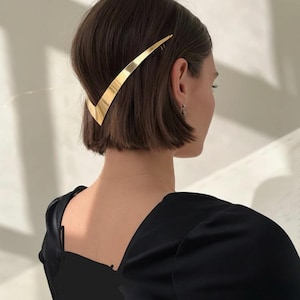 Könnte beinhalten: Ein goldfarbener Haarklammer in V-Form wird in dunklem braunem Haar getragen. Die Person trägt ein schwarzes Oberteil. Das Haaraccessoire ist ein schlichtes, elegantes Design.