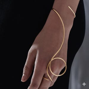 May include: A gold-coloured, minimalist bracelet and ring set. The bracelet curves around the wrist and connects to a ring via a delicate, flowing line. The ring is a simple band. The jewellery is displayed against a dark background.