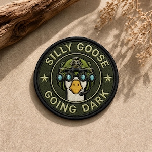 May include: A circular embroidered patch featuring a white goose wearing a green tactical helmet with night vision goggles. The text SILLY GOOSE GOING DARK is stitched in a circular border around the goose, accented by two small stars on the sides.