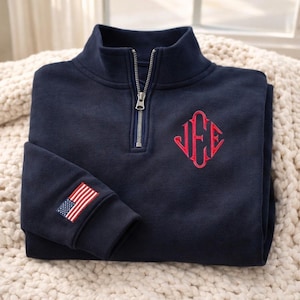 Könnte beinhalten: Marineblaues Sweatshirt mit Viertelreißverschluss, rotem Monogramm und einem amerikanischen Flaggenaufnäher am Ärmel. Das Sweatshirt ist auf einer cremefarbenen, strukturierten Oberfläche gefaltet. Der Reißverschluss ist silberfarben.