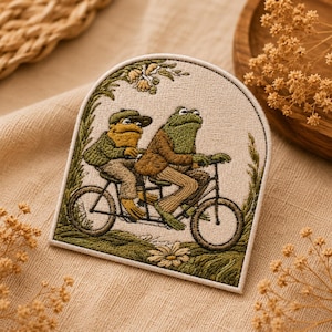 May include: An arched embroidered patch featuring two green frogs riding a tandem bicycle. The frogs wear brown and green clothing, including a cap. The scene is framed by green foliage and a flower at the bottom, all set against a cream background.