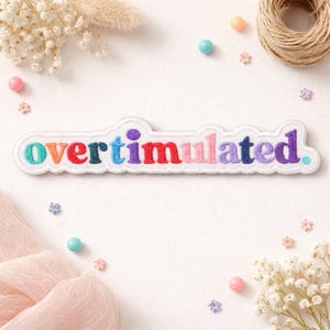 May include: A white embroidered patch featuring the word "OVERSTIMULATED" in a rainbow of colours. The patch is edged in white and is set amongst decorative items including flowers, beads, and twine.