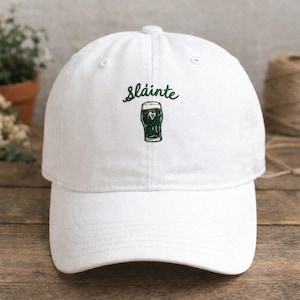 May include: White baseball cap with the word "Sl&aacute;inte" and a green graphic of a pint of stout. The cap has a curved brim and a button on top. The design is embroidered on the front.