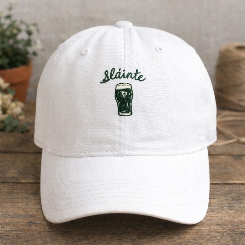 May include: White baseball cap with the word "Sl&aacute;inte" and a green graphic of a pint of stout. The cap has a curved brim and a button on top. The design is embroidered on the front.