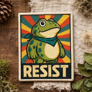 May include: Embroidered patch with a cartoon frog wearing a teal bandana, set against a retro-style background of orange, yellow, and green rays. The word "RESIST" is displayed in bold, gold lettering.