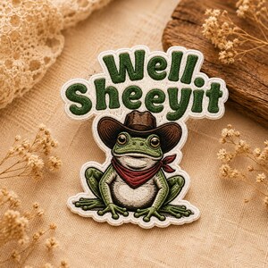 Puede incluir: Un parche bordado que muestra una rana verde con un sombrero de vaquero marrón y un pañuelo rojo alrededor del cuello. Sobre la rana, el texto Well. SHEEYIT está bordado en hilo verde oscuro con un borde blanco sobre un fondo claro.