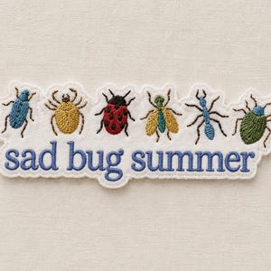Peut inclure: Écusson brodé avec six insectes colorés : un scarabée bleu, une araignée jaune, une coccinelle rouge, un insecte vert, une fourmi bleue et un scarabée vert. Le texte "sad bug summer" est brodé en bleu en dessous.