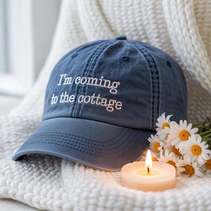 May include: A blue baseball cap with the embroidered text "I'm coming to the cottage." The cap is resting on a white knitted blanket, next to a lit candle and a bouquet of daisies.