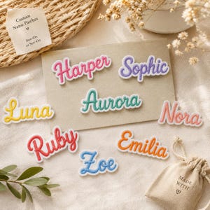 Könnte beinhalten: Eine Auswahl an Namensaufnähern in verschiedenen Farben und Schriftarten, darunter Harper, Sophie und Luna. Die Aufnäher haben einen weißen Rand. Das Bild zeigt auch einen kleinen Beutel mit dem Text "MADE WITH" und einem Herzsymbol.
