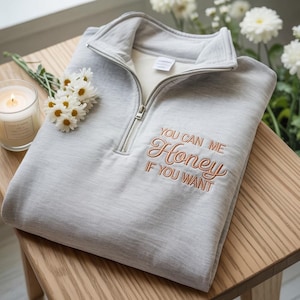 Może przedstawiać: Jasnoszara bluza z zamkiem błyskawicznym do połowy szyi z wyhaftowanym napisem "YOU CAN ME Honey IF YOU WANT". Bluza jest złożona na drewnianej powierzchni, z zapaloną świecą i kwiatami w pobliżu.