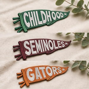 Pode incluir: Três patches bordados em forma de flâmula em verde, bordô e laranja. O patch verde diz "CHILDHOOD", o patch bordô diz "SEMINOLES" e o patch laranja diz "GATORS". Cada patch tem uma estrela e franjas decorativas.